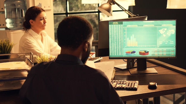 A person sitting at a desk, working on a business plan, with various charts and graphs displayed on a monitor in the background. The scene conveys focus and determination.