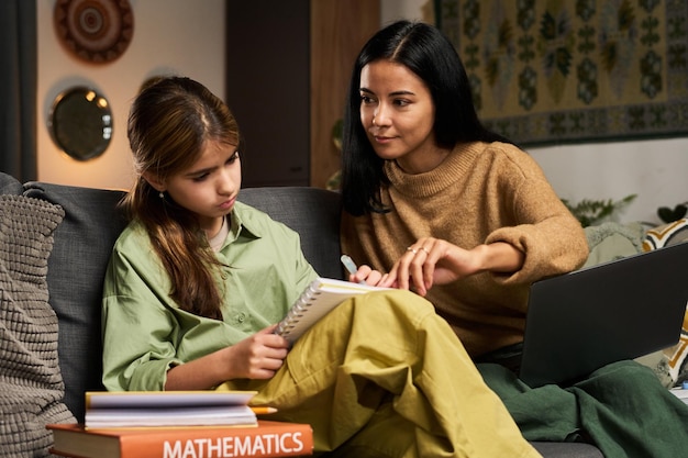 A female educator with a friendly smile is teaching mathematics to a young student via a video call. They are both using digital tablets, and there are math textbooks and notes surrounding them on the desk.